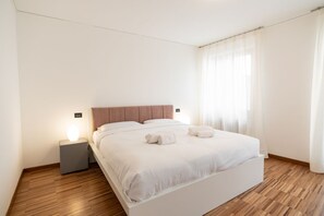 Un dormitorio moderno con suelo de madera, una cama grande con ropa de cama blanca, dos mesitas de noche con lámparas y cortinas translúcidas en la ventana.