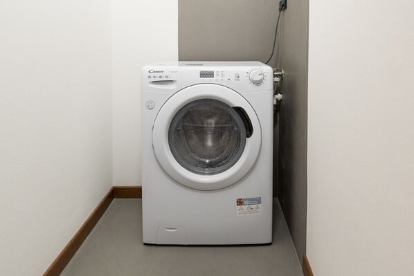 A white Candy brand front-loading washing machine in a corner with a wooden floor and a plain wall.