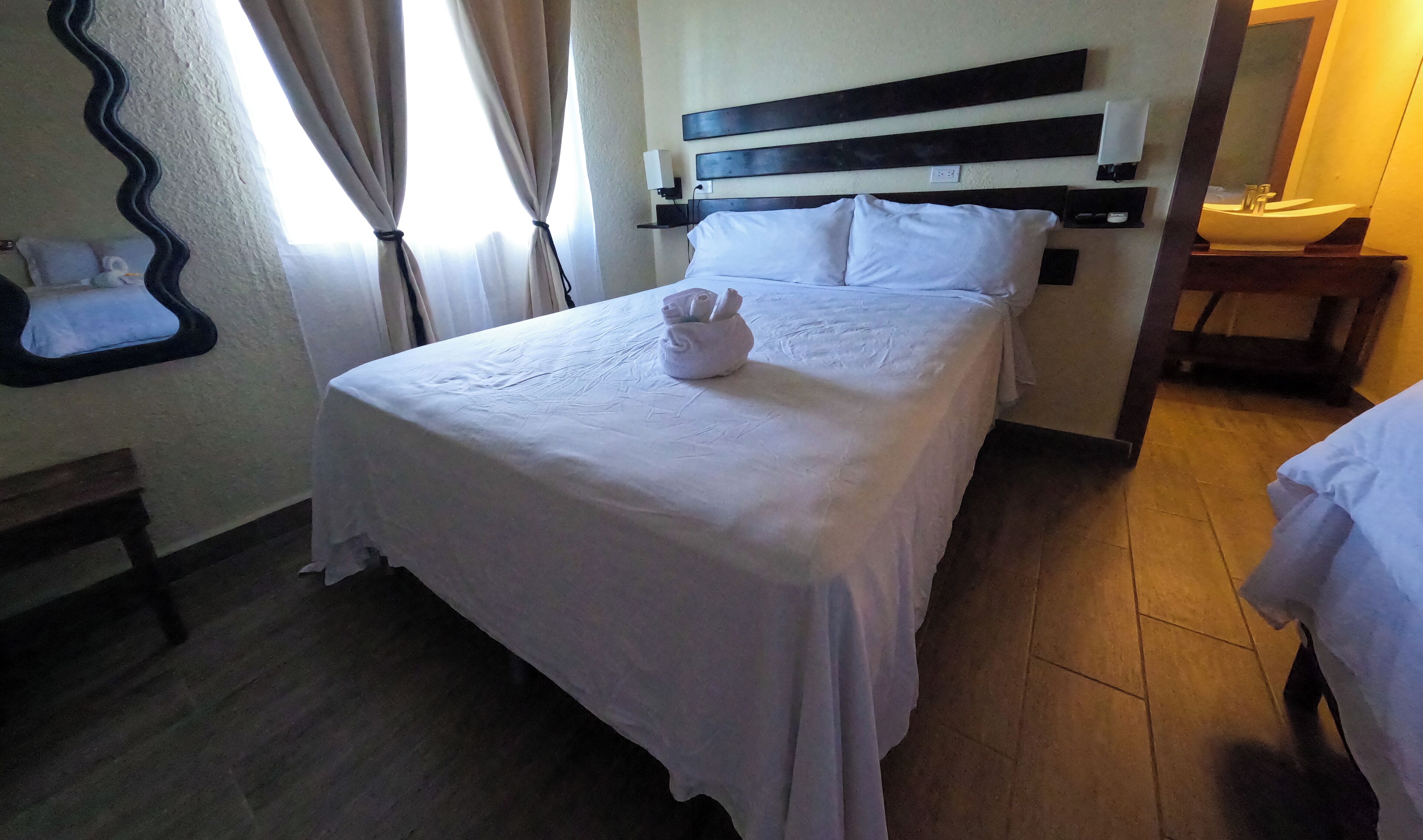 Una cama bien hecha con una colcha blanca, una toalla pequeña sobre la cama y una mesita de noche de madera con un espejo y una pequeña mesa.