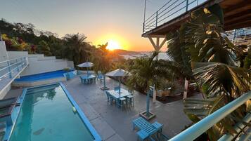 A pool area with a view of the sunset over the water, surrounded by palm trees and outdoor seating.