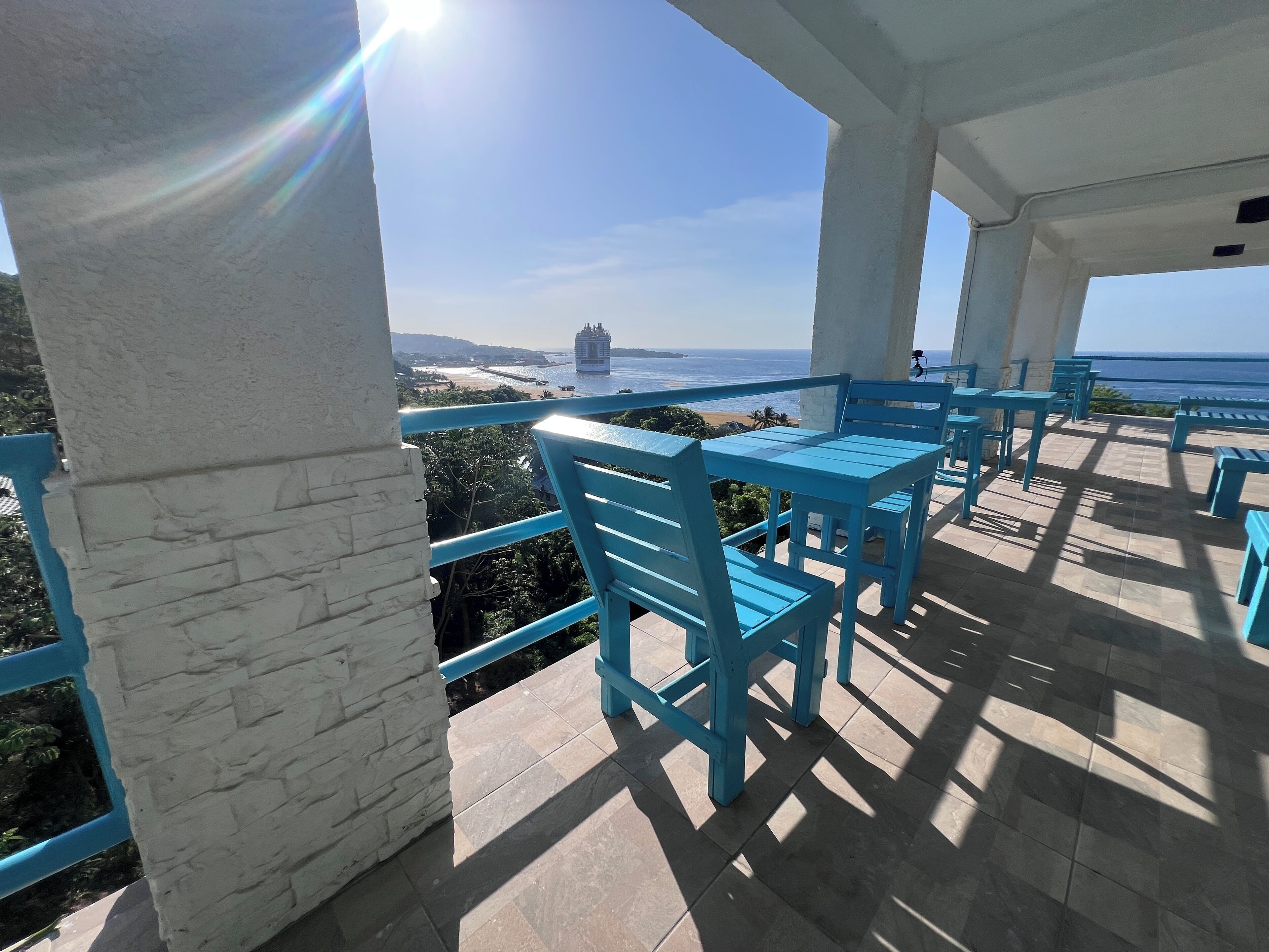 A covered outdoor seating area with blue chairs and tables, overlooking a beach and ocean.