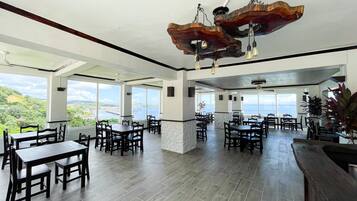 A spacious dining area with high ceilings, large windows offering a view of the sea, and a unique wooden ceiling light fixture.