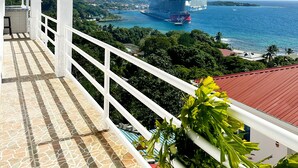 Un balcone con pavimento piastrellato e ringhiera bianca con vista su una nave da crociera e una cittadina costiera.
