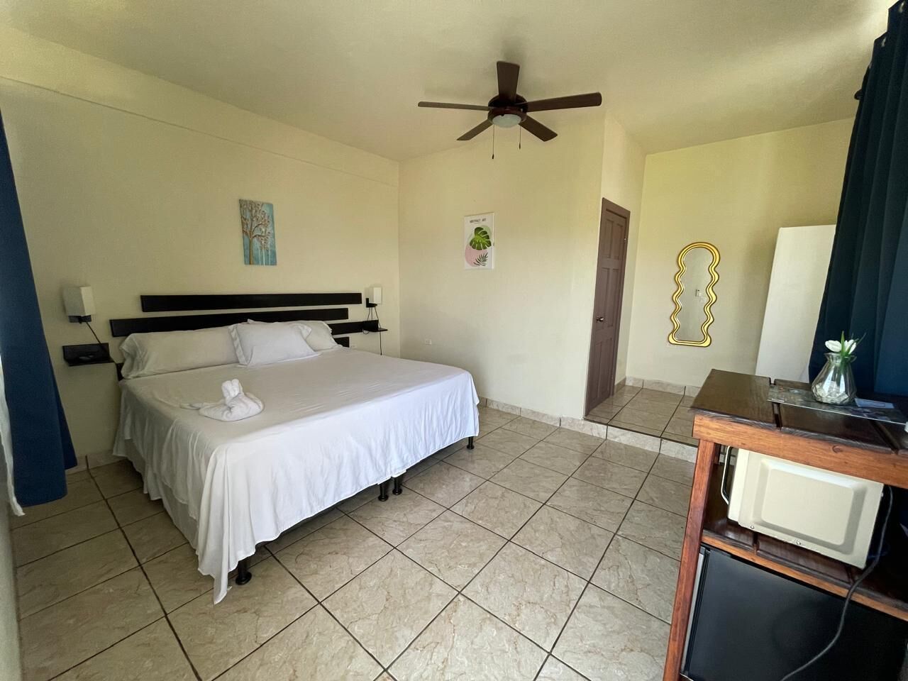 A hotel room with a bed, a ceiling fan, a small table with a vase, and a door leading to another room.