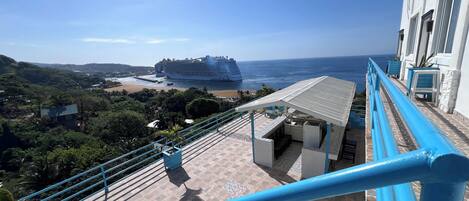 A view from a balcony overlooking a beach with a cruise ship in the distance, a patio area with outdoor seating, and a building with a sign reading "OCEAN VID HOTEL RESTAURANT".