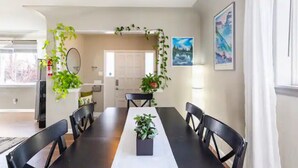 A dining area with a long table, black chairs, and potted plants. There is a white door in the background, and a window with curtains.