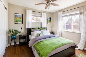 Une chambre à coucher comprenant un lit, une table de chevet, une lampe, une fenêtre avec des rideaux et un ventilateur de plafond.