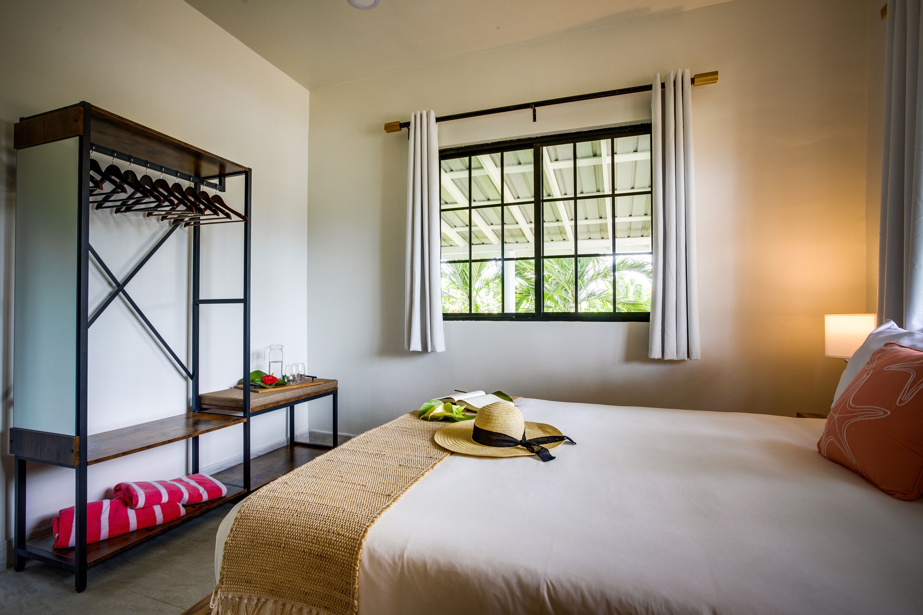 Una habitación de hotel con una cama, un perchero, una ventana con cortinas y vistas al exterior con vegetación.