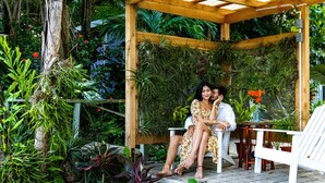 Una pérgola de madera con suelo de baldosas, rodeada de abundante vegetación y plantas en macetas.