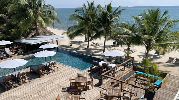 Terraza de madera con sillones de descanso y sombrillas, con vista a una piscina y una playa.