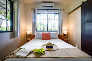 Una cama bien hecha con un libro, un sombrero y una planta sobre un tapete tejido. Hay dos lámparas de noche, un cojín con forma de estrella y una ventana con cortinas.