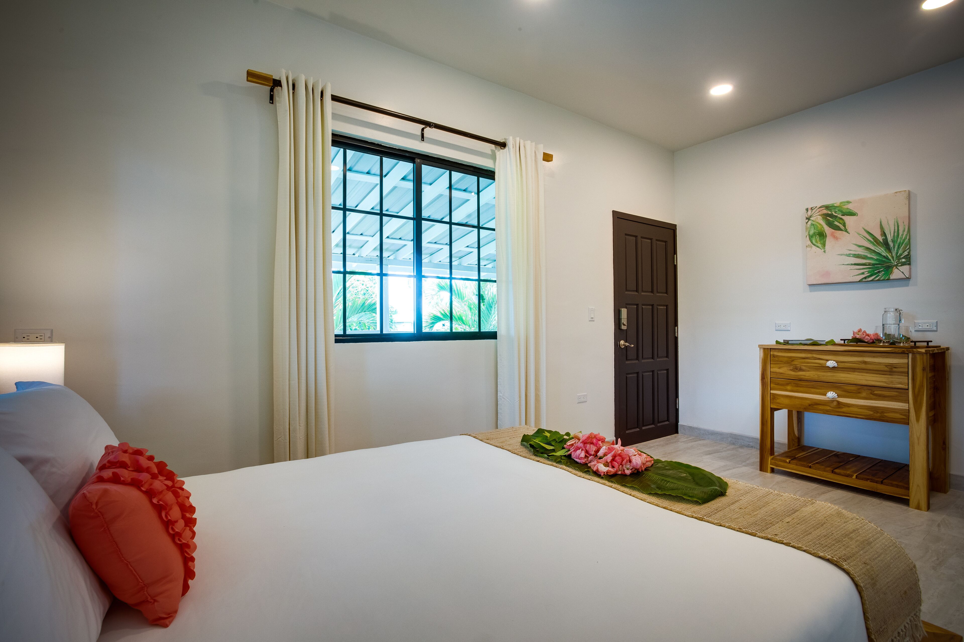A hotel room with a bed, a wooden nightstand, a window with curtains, a door, and a wall-mounted picture.