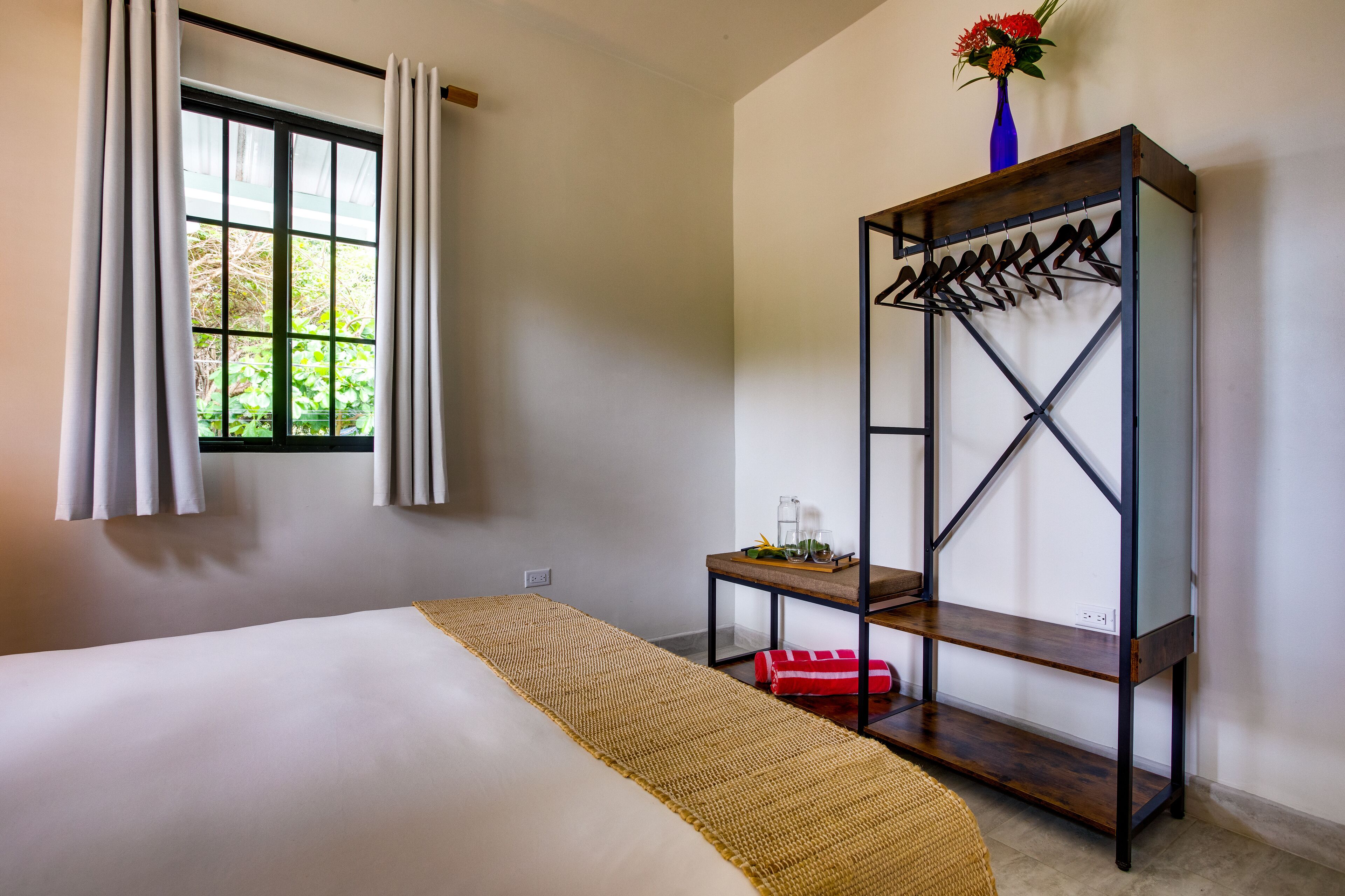 Un dormitorio con cama, ventana con cortinas, estante de madera con un florero y una mesita con vaso y planta.