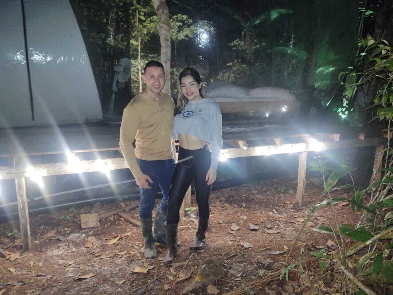 Two people standing outdoors at night with a wooden structure and string lights in the background.