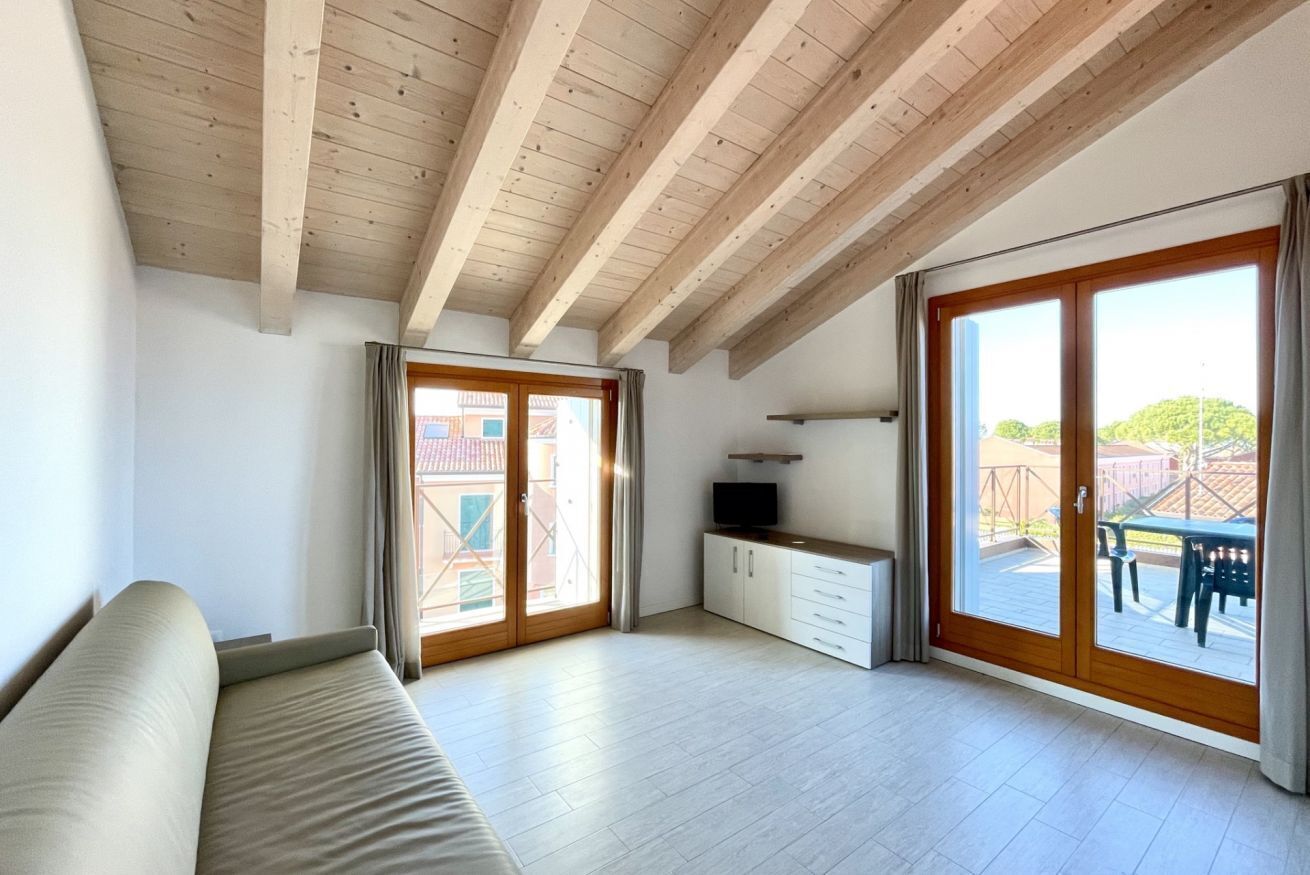 A spacious living area with a wooden ceiling, a sofa, a TV on a cabinet, and large sliding glass doors leading to an outdoor patio.