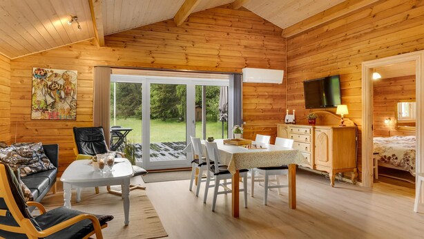 Interior acogedor de una cabaña con comedor, sala de estar con sofá y sillones, zona de cocina con televisor y dormitorio con cama.