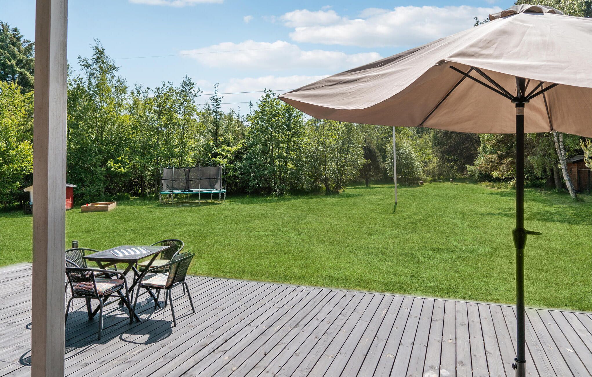 Eine Holzterrasse mit Tisch und Stühlen, ein überdachter Sonnenschirm und ein Rasen mit Bäumen sowie eine Bank im Hintergrund.