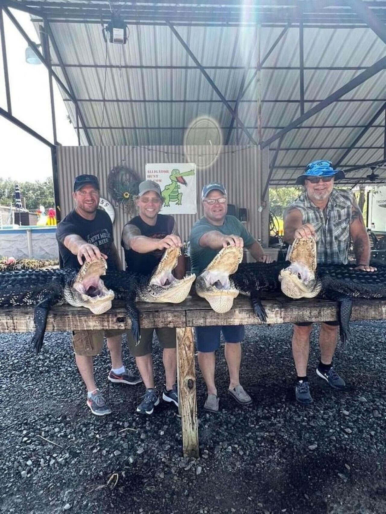 Four people sitting on a bench under a metal roof, each holding a large alligator head.
