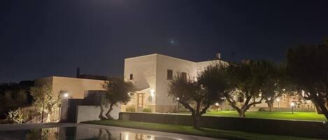 Una vista nocturna de un edificio moderno con piscina, rodeado de árboles e iluminado por farolas.