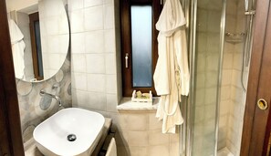 Baño con lavamanos blanco, espejo, cabina de ducha de vidrio y ventana con toallero.
