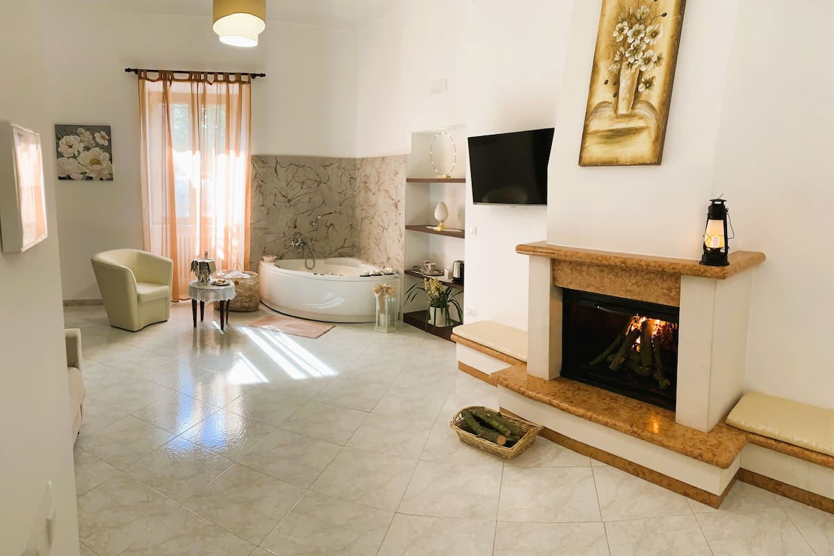 A spacious living room with a fireplace, a large window with curtains, a sofa, a coffee table, and a bathtub.