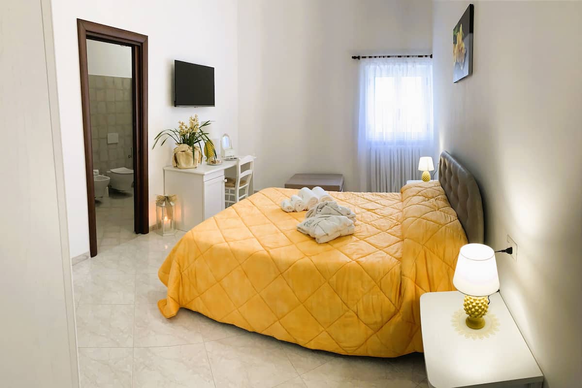 A hotel room with a bed, bedside table with a lamp, a desk with a plant, a mirror, and a TV on the wall.