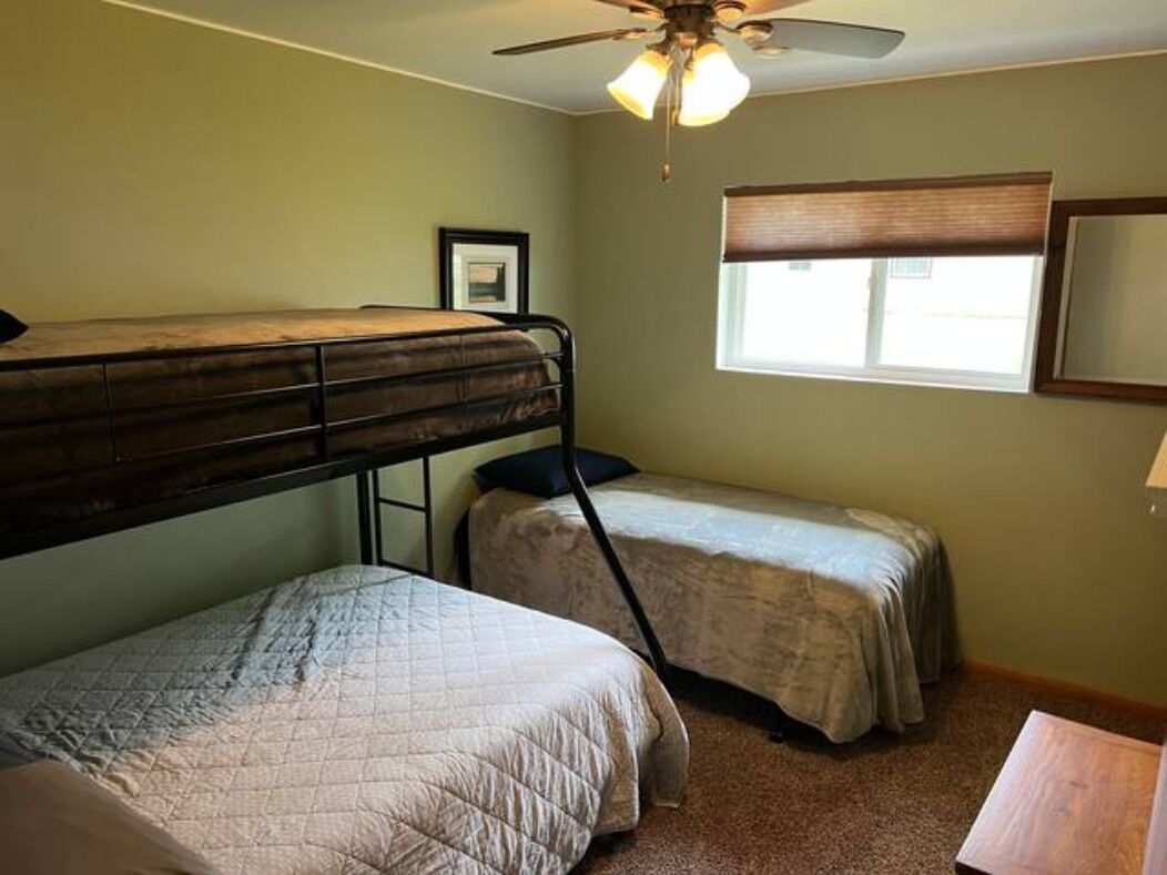 A dormitory room with bunk beds, a ceiling fan, a window with blinds, and a framed picture on the wall.
