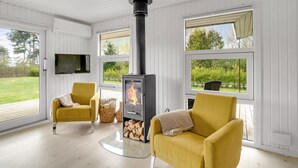 Ein Zimmer mit Kamin, zwei gelbe Sessel, einem Fernseher und großen Fenstern mit Blick auf Grünflächen.