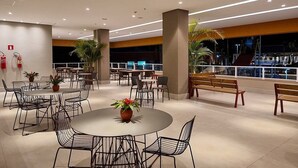 Un comedor moderno con mesas redondas y sillas de metal, rodeado de pilares grandes y plantas en macetas.