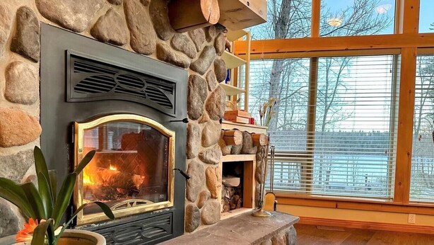 Une cheminée en pierre avec un feu de bois, une plante en pot et une vue sur un lac, visible à travers de grandes fenêtres.