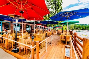 An outdoor dining area with wooden tables and chairs, colorful umbrellas, and a wooden deck overlooking a beach. - Tahoe Views & Tahoe Park Access (Tahoe City)
