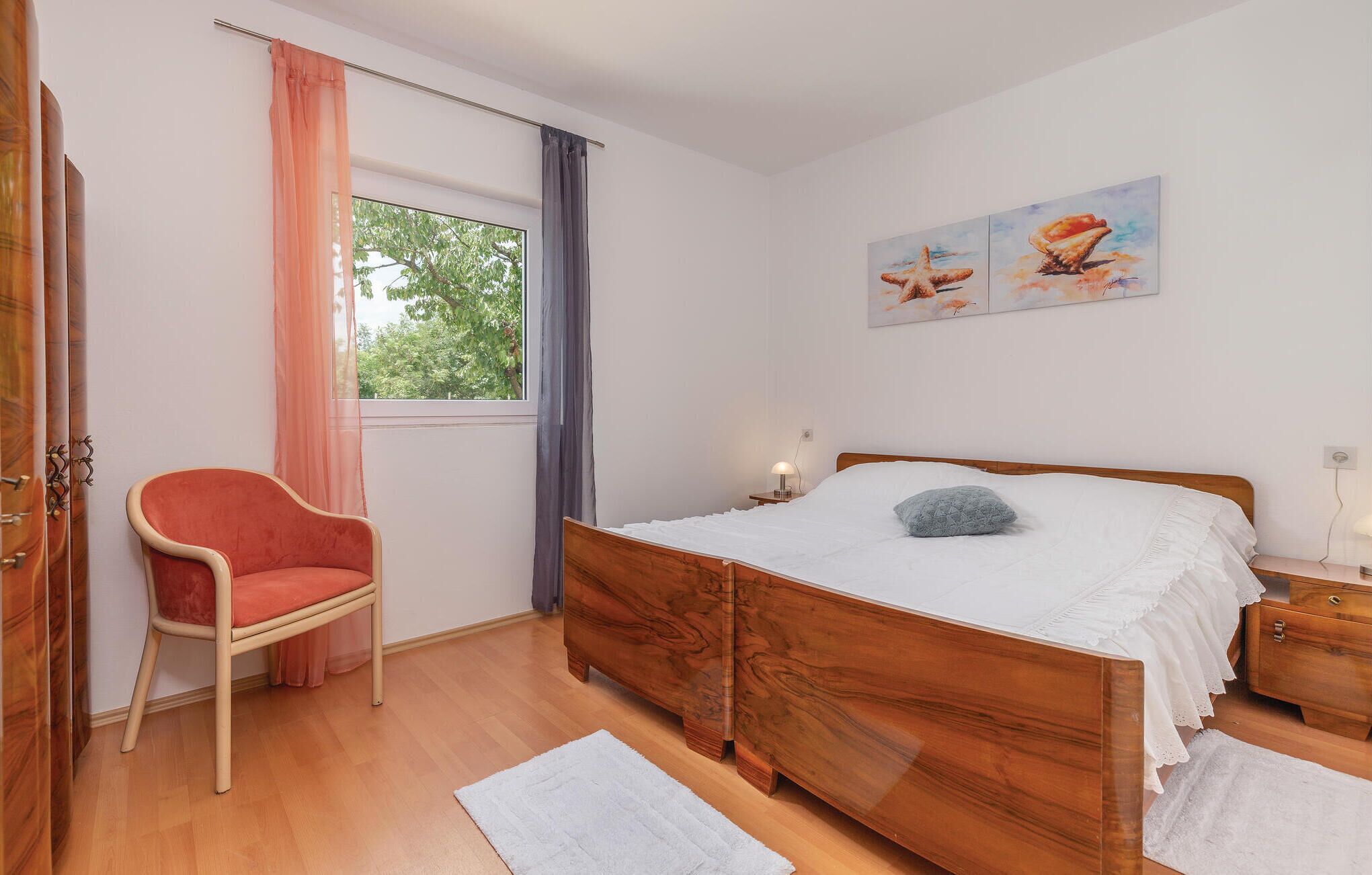 Een slaapkamer met een houten bed, een stoel, een raam met gordijnen en kunst aan de muur.