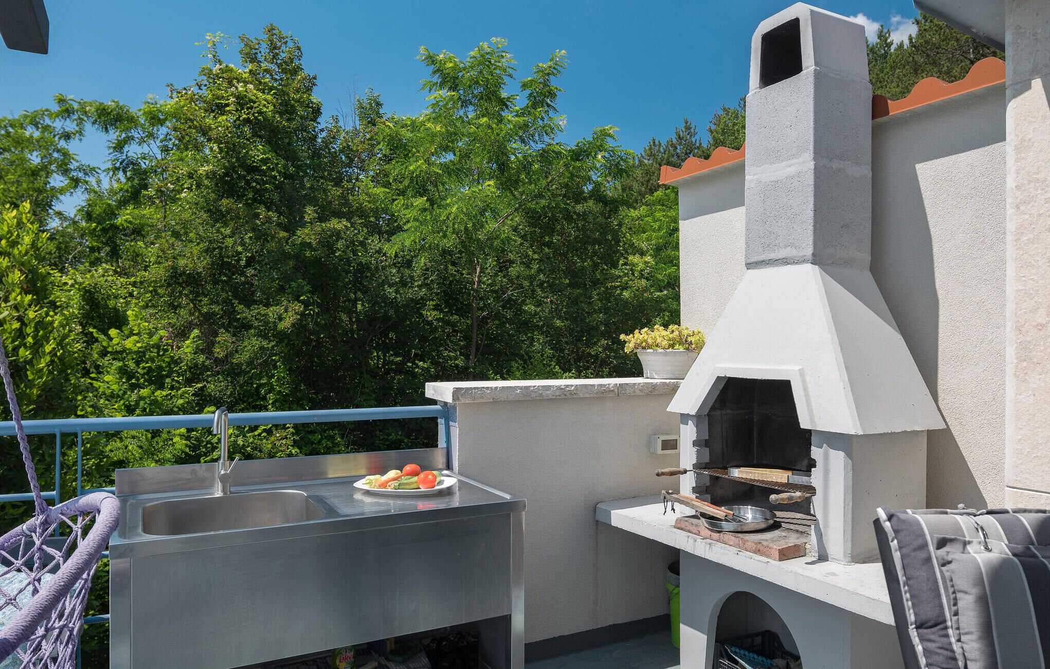 An outdoor kitchen area with a grill and a sink, surrounded by greenery.