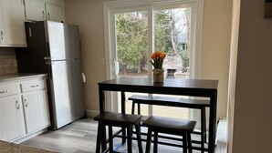 A kitchen with white cabinets, a black countertop, a stainless steel refrigerator, and a window with a view of trees and a stone fireplace. - Narragansett- 1 block to Narrow River! (Narragansett)