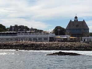 A coastal building with a prominent tower, a rocky shoreline, and a pier with people.