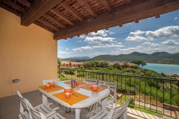 A balcony with a table set for two, overlooking a coastal landscape with mountains and a body of water.