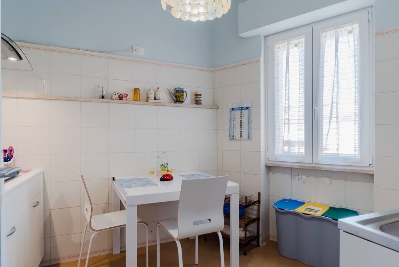 Eine Küche mit weißen Schränken, einem weißen Tisch, Stühlen, einem Fenster mit Jalousien und einem Kalender an der Wand.