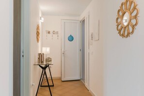 Un couloir avec une porte, une petite table sur laquelle sont posés des livres, un miroir fixé au mur et un élément décoratif mural.