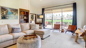 Ein geräumiges Wohnzimmer mit einem Ecksofa, einem Hocker und einem Essbereich mit Stühlen. Ein großes Fenster bietet Blick auf einen Golfplatz.
