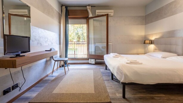A modern hotel room with a bed, a desk with a TV, a chair, a large window, and a balcony. - Appartamento a 200 metri dall\'Arena con 3 camere da letto e terrazzo (Verona)