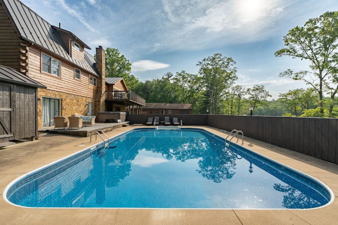 A rectangular swimming pool with a wooden deck surrounding it, a covered seating area with chairs, and a stone building with a metal roof in the background.