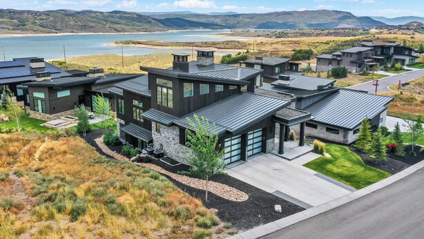 Una zona residencial moderna con casas que tienen ventanas amplias y techos oscuros, rodeada por un paisaje natural con un lago y montañas al fondo.