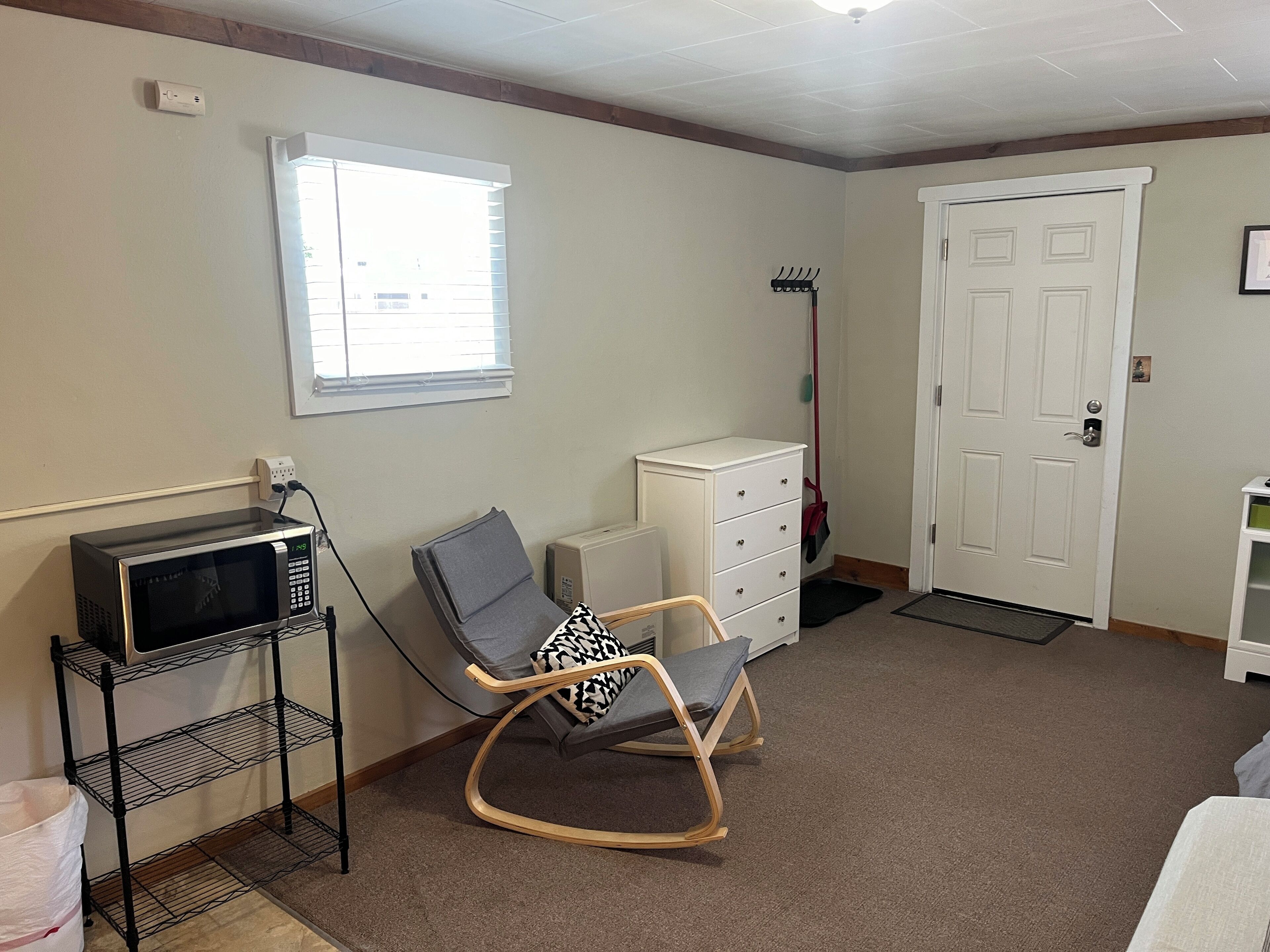 Une pièce avec un fauteuil à bascule, une commode blanche, un four à micro-ondes posé sur une étagère, une porte et une fenêtre avec des stores.