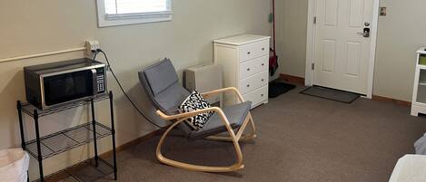 Une pièce avec un fauteuil à bascule, une commode blanche, un four à micro-ondes posé sur une étagère, une porte et une fenêtre avec des stores.