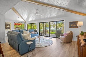 A spacious living room with a high ceiling, a ceiling fan, and large sliding glass doors leading to an outdoor area with a boat and greenery. - Nirvana:Pool & Hot Tub, Dock, On Canal with Gorgeous Water Views by Anna Maria Life Vacation Rentals (Anna Maria)