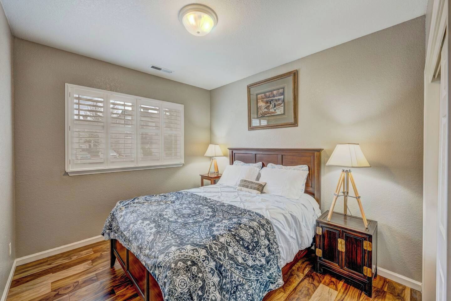 A bedroom with a bed, two bedside lamps, a ceiling light, a window with blinds, and a framed picture on the wall.