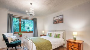 Une chambre à coucher comprenant un lit, une chaise, une table de chevet, une lampe, une fenêtre avec des rideaux, un lustre et un tableau accroché au mur.