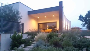 Een modern huis met een overdekte buitenruimte, omgeven door een tuin met diverse planten en stenen.