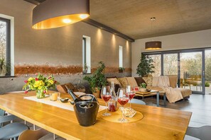 Un espace repas moderne avec une table en bois, des verres à vin et une bougie. On aperçoit un canapé et un fauteuil au fond.