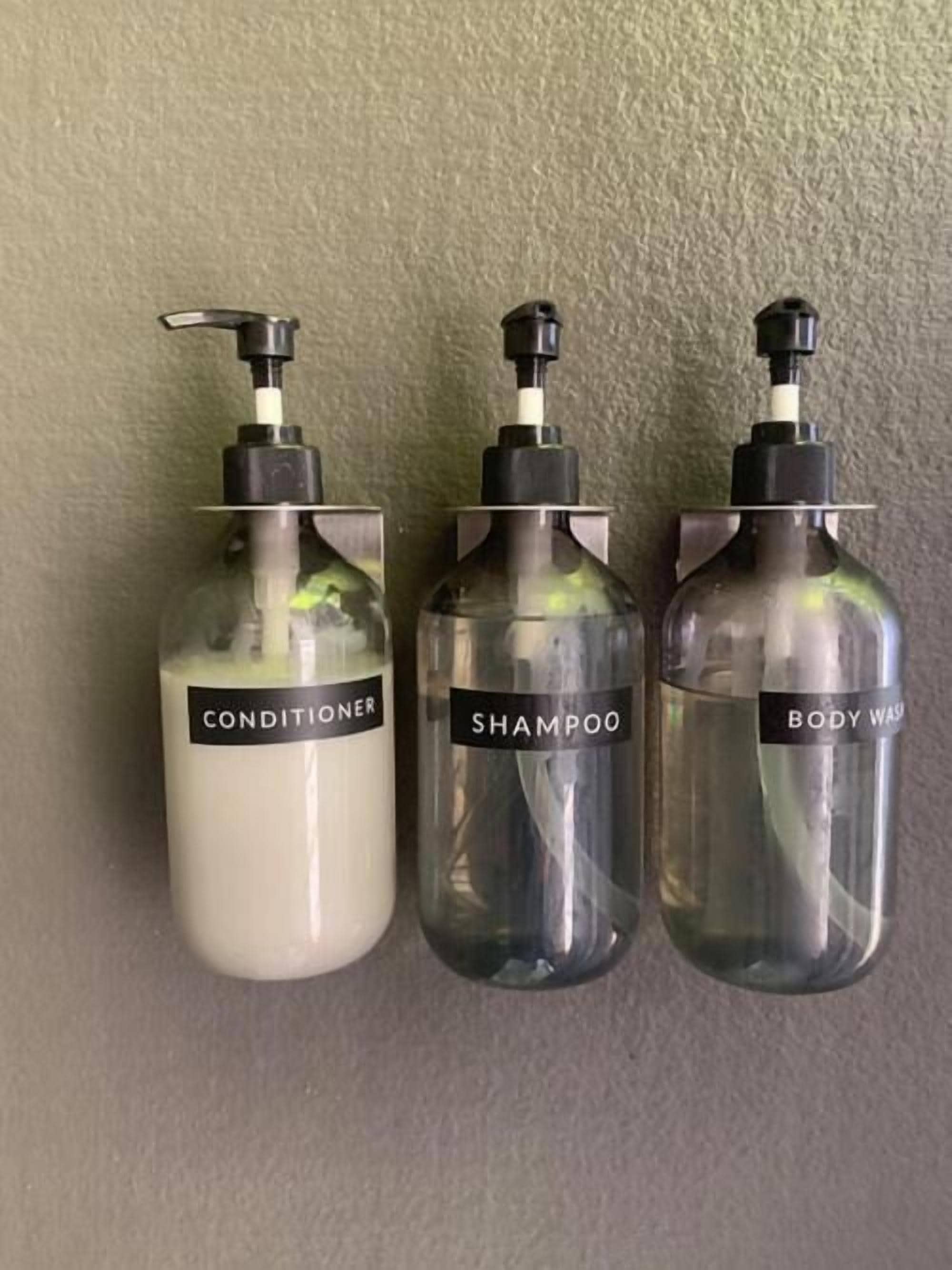 Three transparent bottles with pumps, labeled 'CONDITIONER,' 'SHAMPOO,' and 'BODY WASH,' against a textured wall.